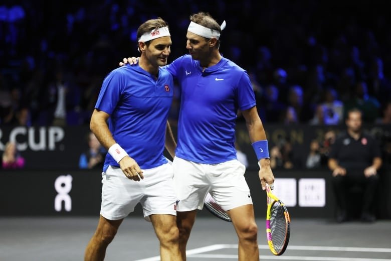 Two cleanshaven tennis players in headbands gather closely on the court, with one putting his arm around the other.