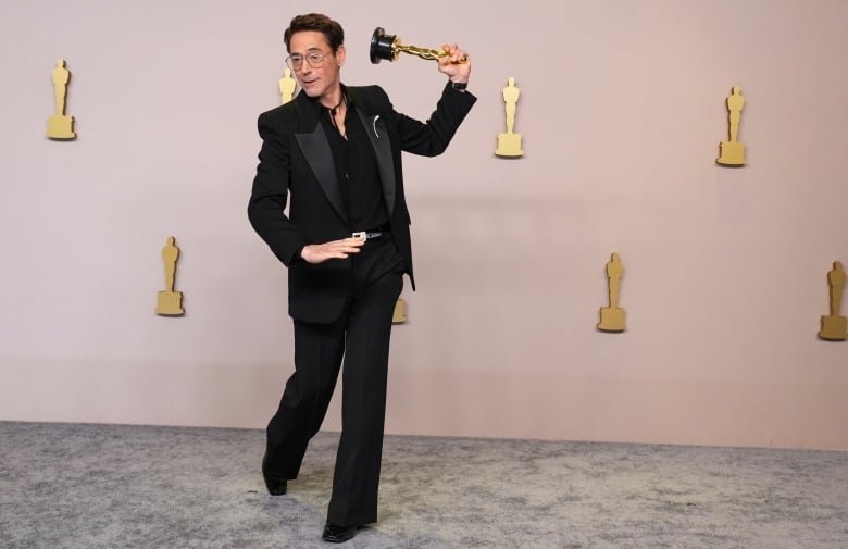 A man is shown on a velvet carpet in a suit. He has one leg kicked bent behind him in a jaunty fashion and is gesturing with his Oscar statue as though he is going to throw it or swing it like a bat or break into a dance.