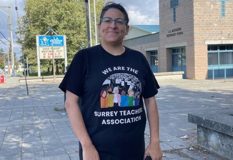 A woman in a black t-shirt with the phrase 'We are the Surrey Teachers' Association' smiles outside, with a high school building seen in the background behind her.