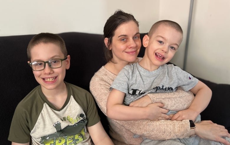 ashley-ricks-with-her-sons A boy with glasses smiles while sitting on a dark couch next to his mother, who holds his younger brother on her lap.