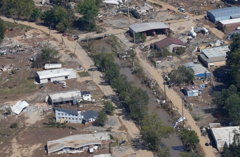 An aerial view of a town shows some smashed buildings or leaning buildings. Others stand intact, but near piles of rubble.