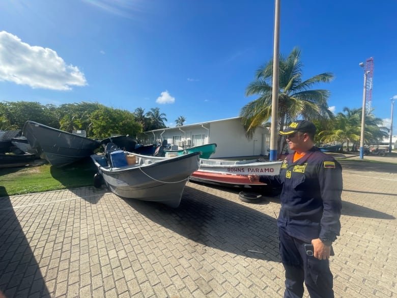 A man in a navy uniform points at several small boats docked on land next to a one-storey building surrounded by palm trees.