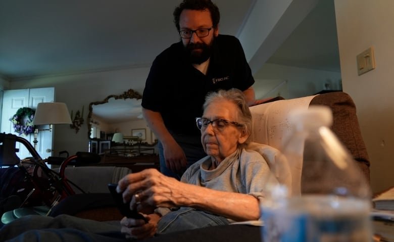 An elderly person sits in a brown leather chair, while a younger man leans over them from the back of the chair. A lamp at the right lights up the otherwise dark room.