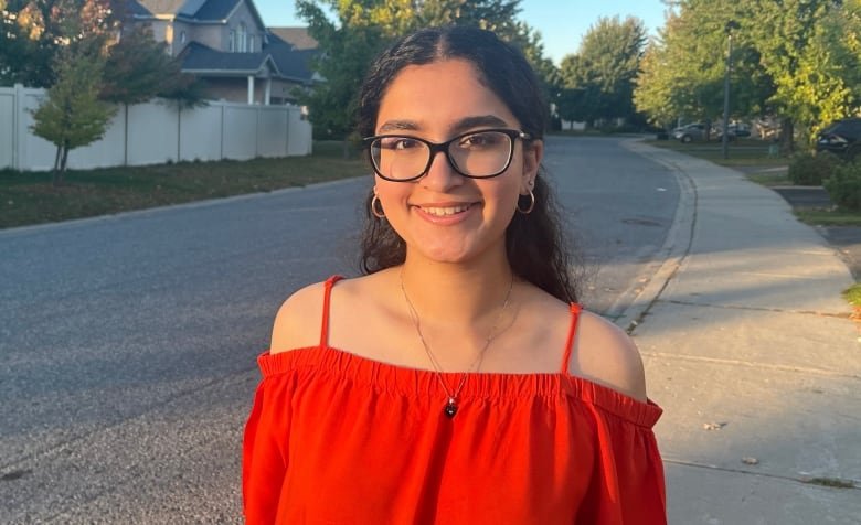 A teen girl wearing a red top and glasses stands smiling outdoors, with a surburban neighbourhood seen behind her.