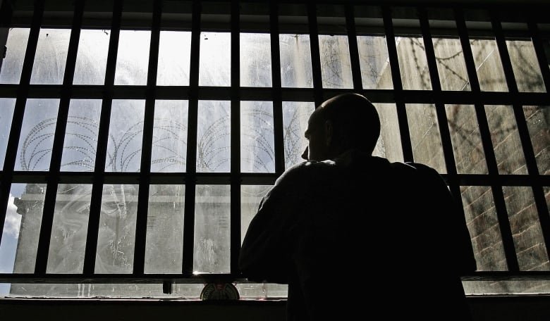 A man looks out a dirty window. Barbed wire is outside the window.