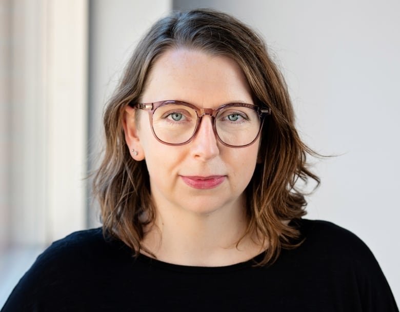 A woman with brown shoulder-length hair and eyeglasses, wearing a black top, looks straight at the camera.