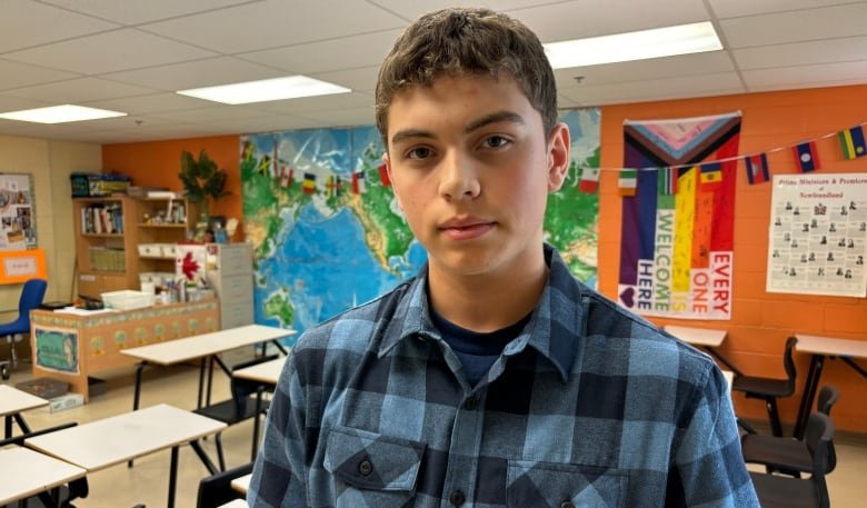 A teen boy in a blue plaid shirt stands in an empty school classroom, with rows of desks, a large map and colourful flags seen behind him.