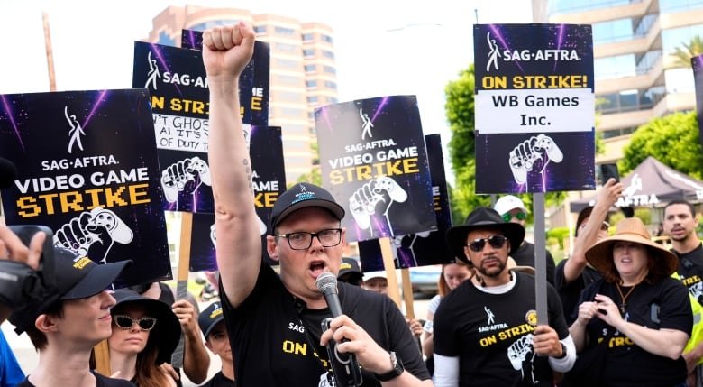 A crowd of people are holding sings that say SAG-AFTRA Video Game Strike. A man in the centre is speaking into a microphone and raising his fist in the air.