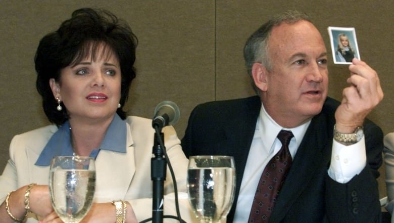 A woman and a man sit at a table with a microphone stand in front of them. The man is looking off to the right side and is holding a small photograph of a young girl up.