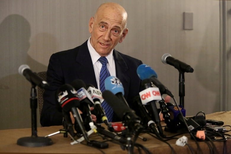 A bald man with looks into the camera surrounded by microphones.