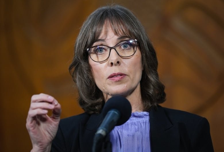 A portrait of a woman in glasses speaking on a microphone.