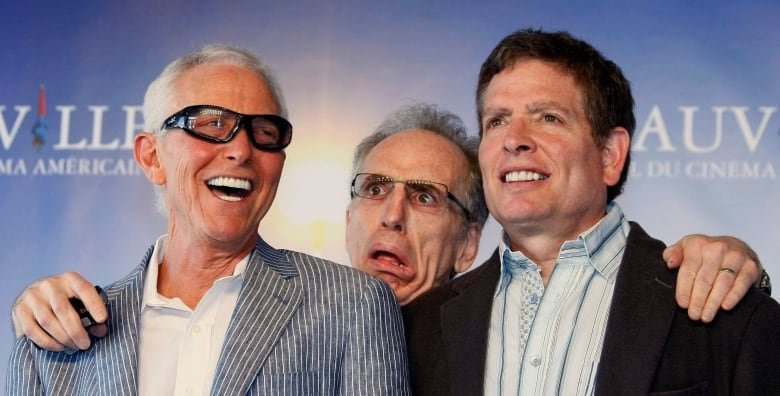 Three men stand in front of a blue backdrop. The man at the left is laughing, the man is in the middle is making a funny face and has his hands on the shoulders of the other two, and the man at the right side is smiling.