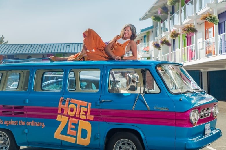 Photo of a woman in orange overalls lounging on a blue camper van. The van has the words Hotel Zed on the side.