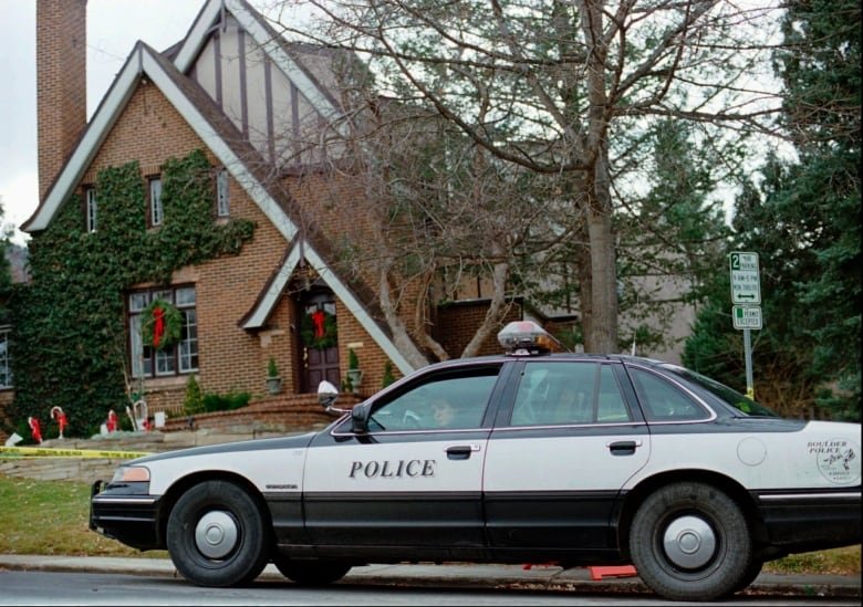 An older model of police car sits outside of a brown house with triangular peaks over the front door and the overall shape of the house.