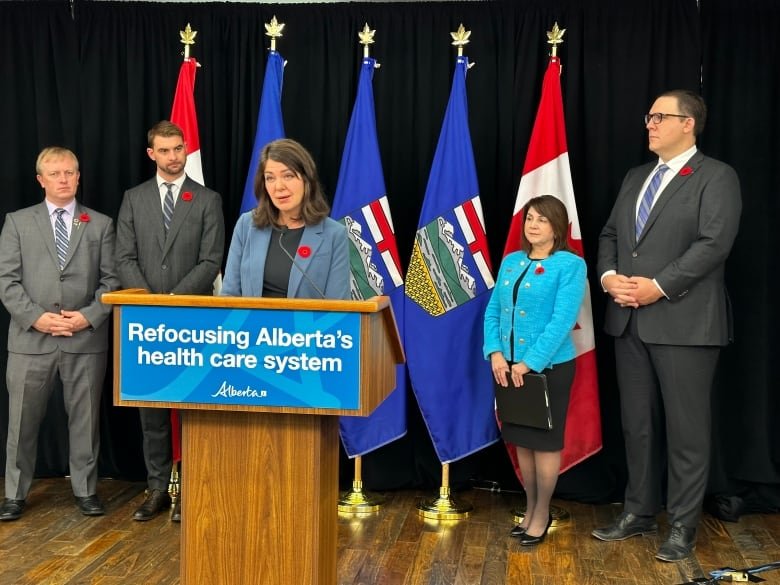 A woman speaks into a microphone at a podium bearing a sign that states Refocusing Alberta's Health Care System. Behind her are three men and one woman, along with a selection of Canada and Alberta flags.