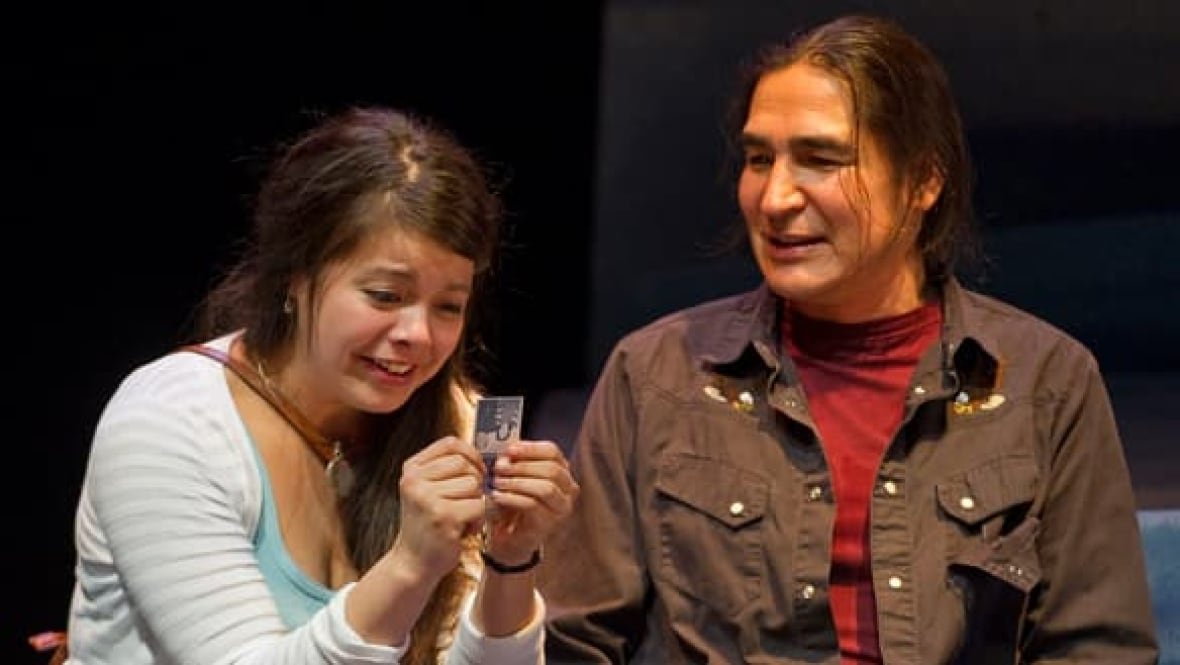 A woman holding a small photo sits beside a man in a scene from a theatre production.