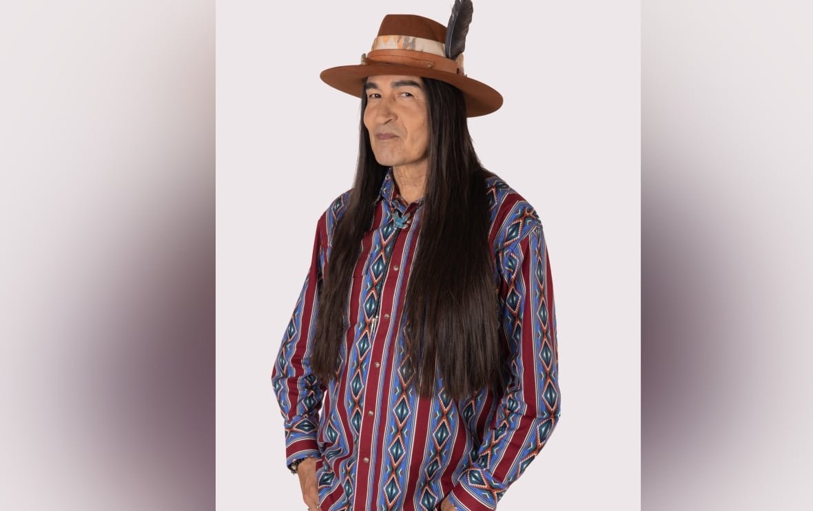 A publicity photo shows a man with long hair wearing a patterned shirt and a hat with a feather in it.