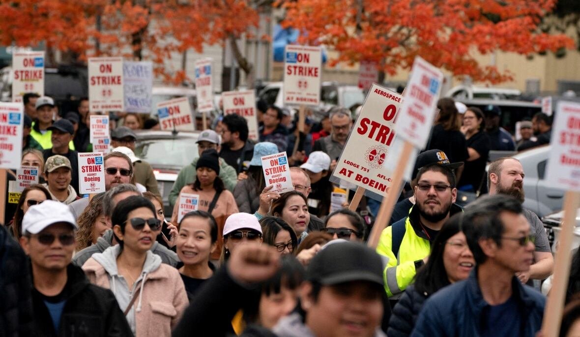 A crowd of people carry signs that say "On Strike". At the top of the image, red fall leaves are visible from a tree in the distance.