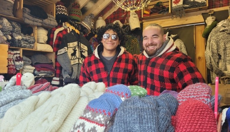 Two men stand in a small shop where mittens and hats are on display.