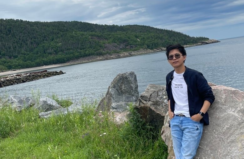 A man wearing jeans, a white t-shirt and a black jacket leans against a large rock. Behind him is a strait of water, a wooded coast, and a cloudy sky.