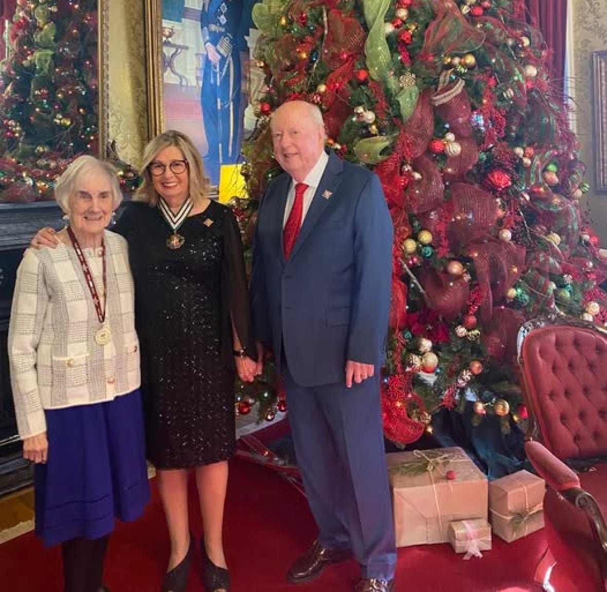 Three standing in front of a tall Christmas tree. one person has a medal around her neck.