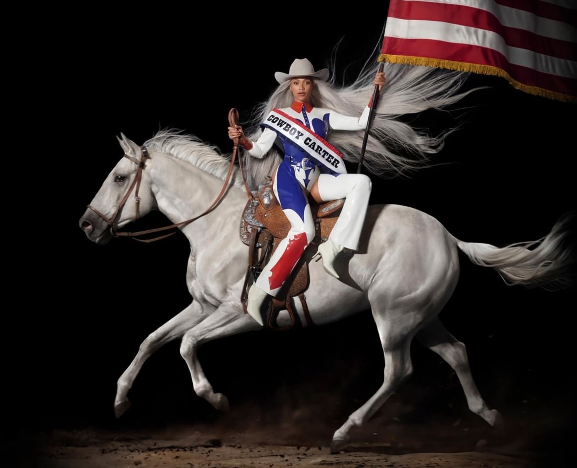 A woman sits sideways on a white horse that is mid-gallop. She is holding an American flag and wearing a sash that says "Cowboy Carter". The background of the image is black.