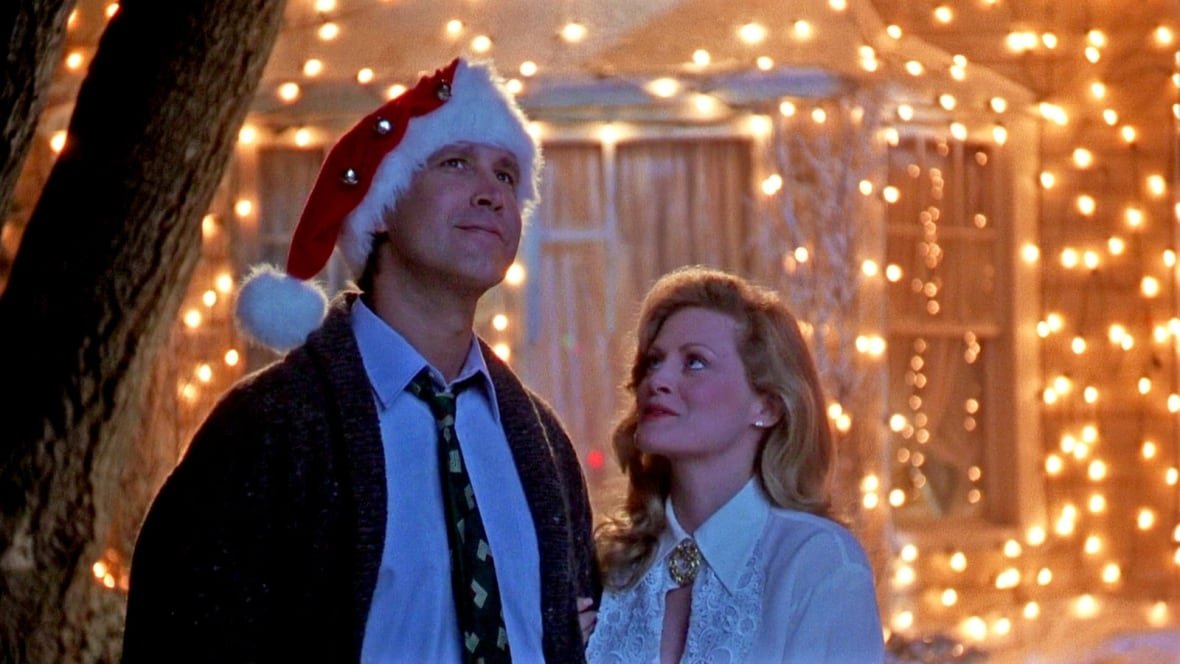 A man in a Santa hat and woman in a blouse stand outside a brightly-lit house.