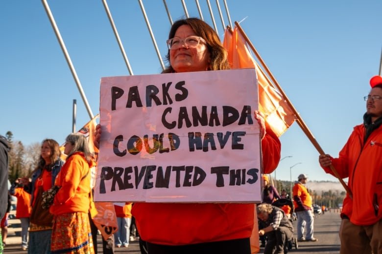 A person is seen holding a sign that says "Parks Canada could have prevented this."