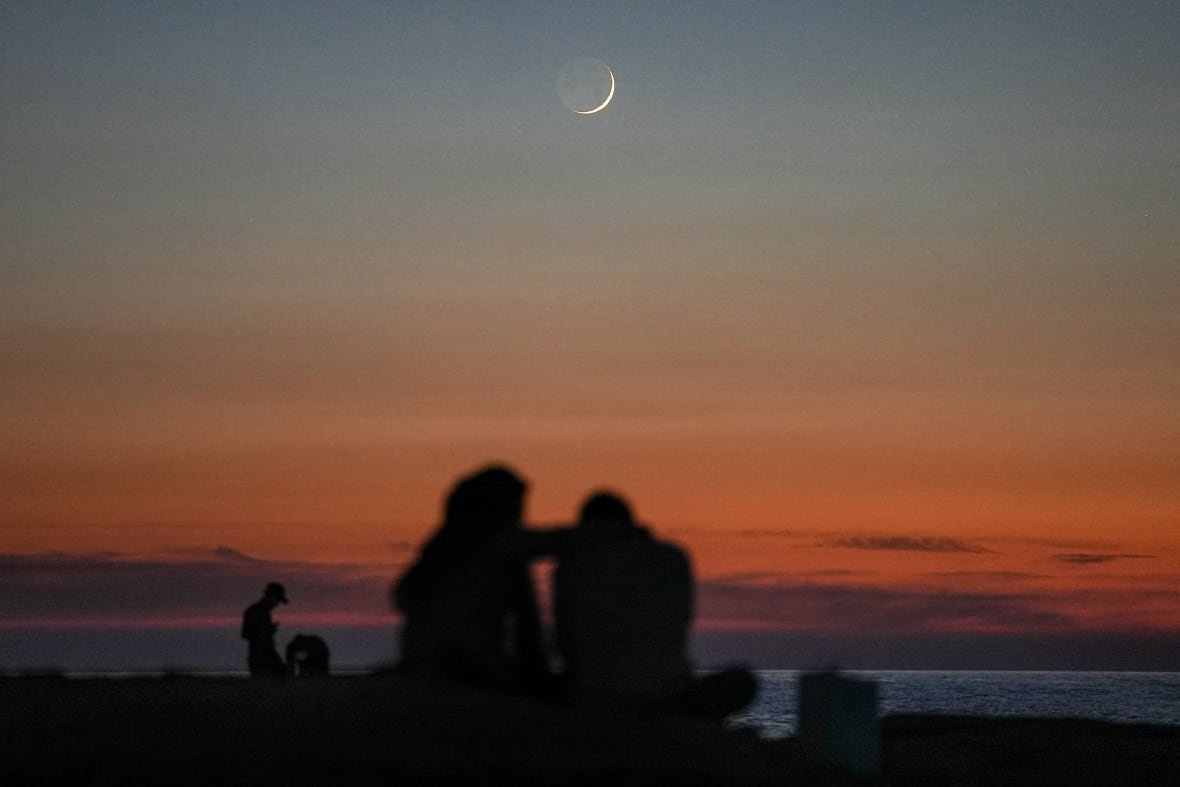 A couple sitting by the sea shore are in silouette against an orange/rust coloured sunset with a tiny sliver of the moon visible high in the sky.