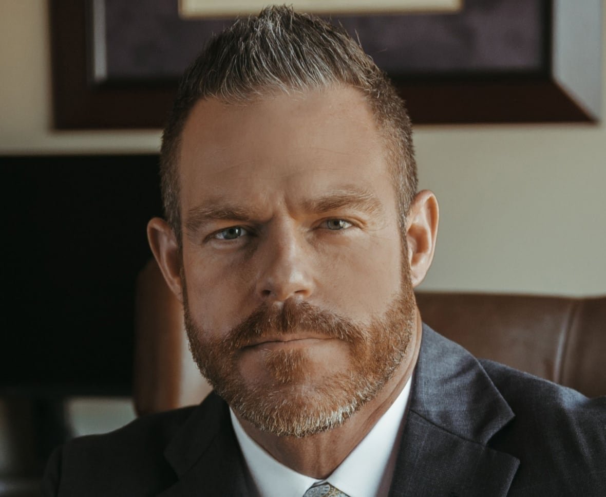 A headshot of a man with short hair and a beard wearing a suit smiles at the camera.