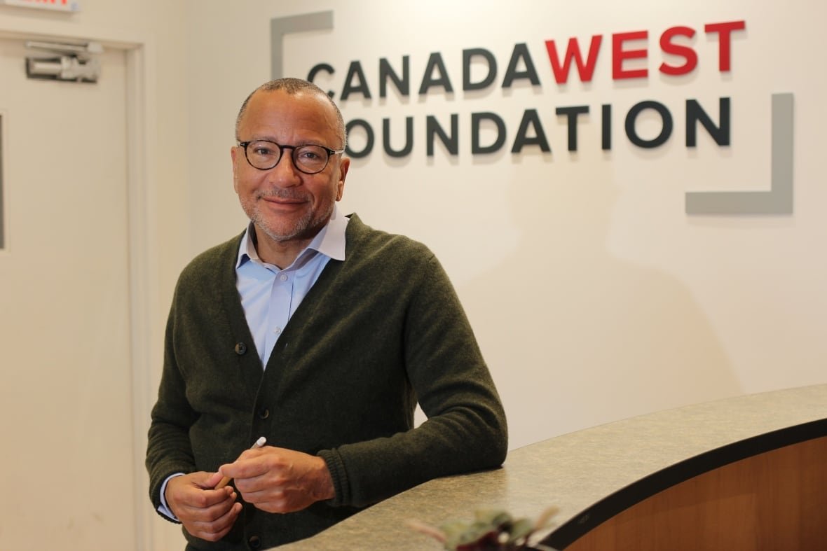 A man wearing glasses, a blue button-down shirt and green cardigan stands in front of a wall bearing the logo of the Canada West Foundation.