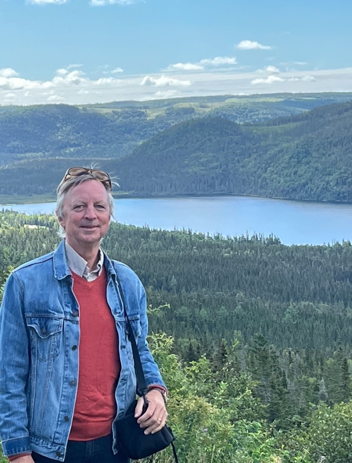 A man standing amid greenery with a lake and hill in the background.