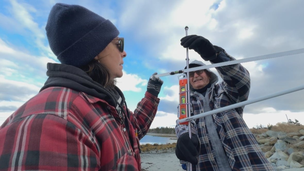 Two people wearing plaid shirts make measurements of a beach using a device made from curtain rods and a level.