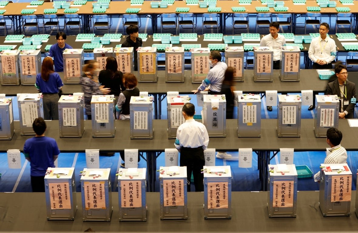 A view from an elevated level shows a room full of boxes for ballots, which contain Asian writing on them. Several people, men and women, are shown in the room.