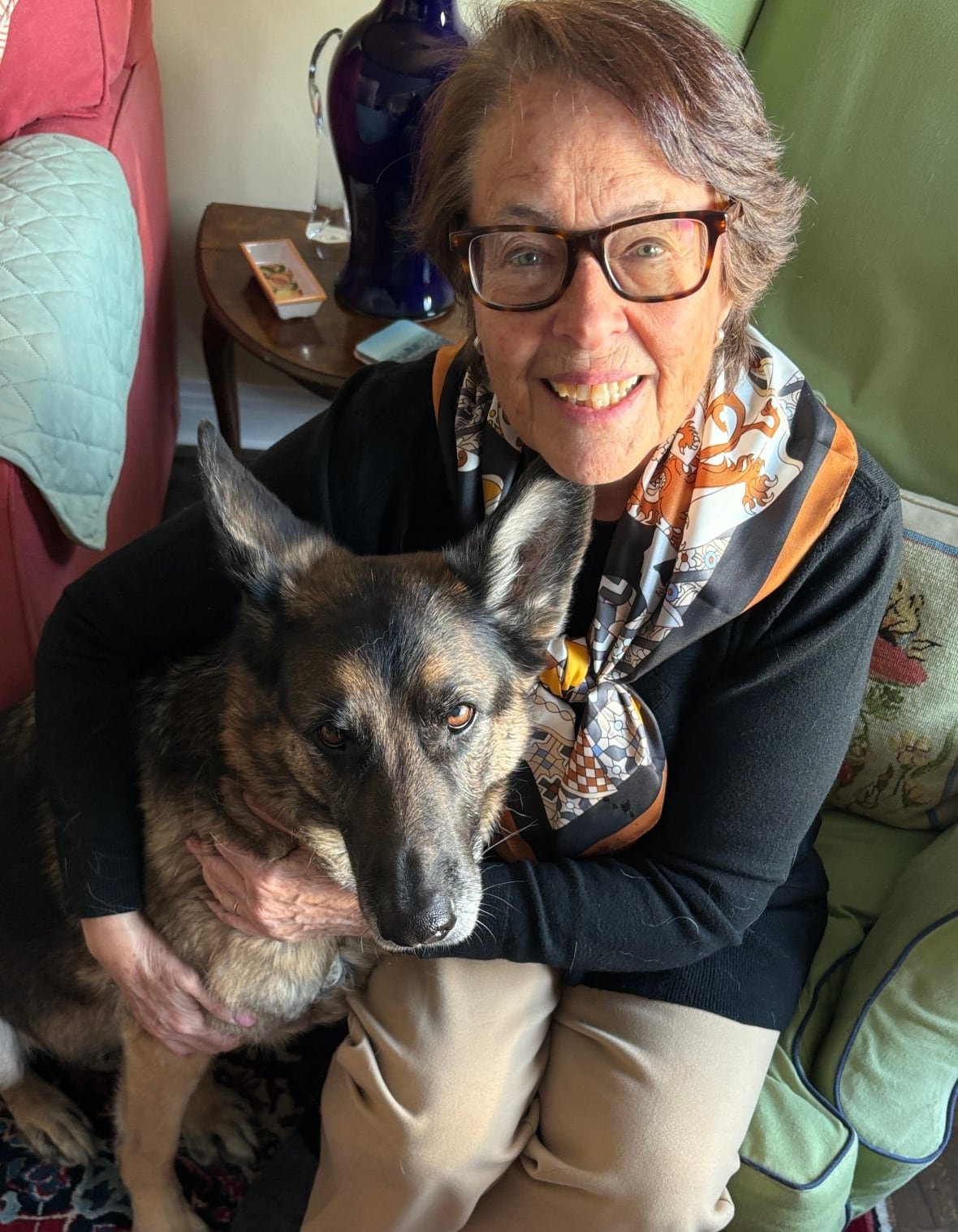 Woman sits on armchair holding her German Shepherd close and looking up at the camera