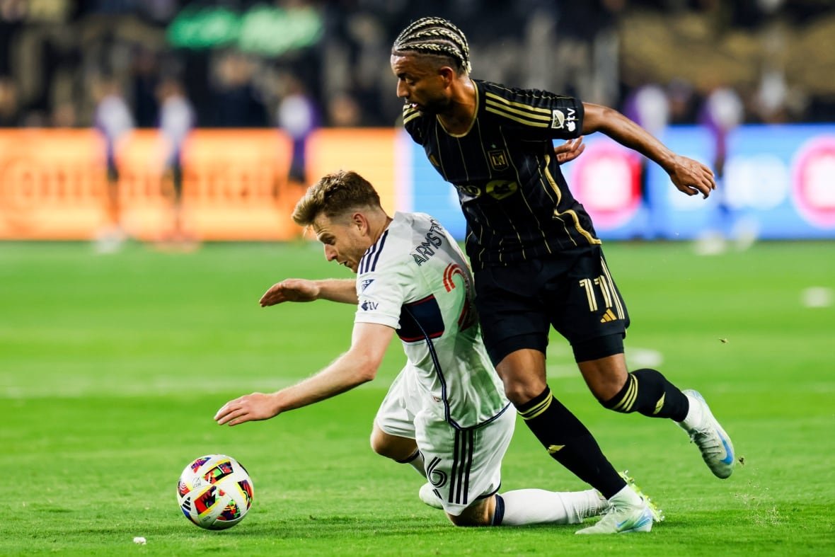 A soccer player falls to the ground near the ball as another player puts on pressure.