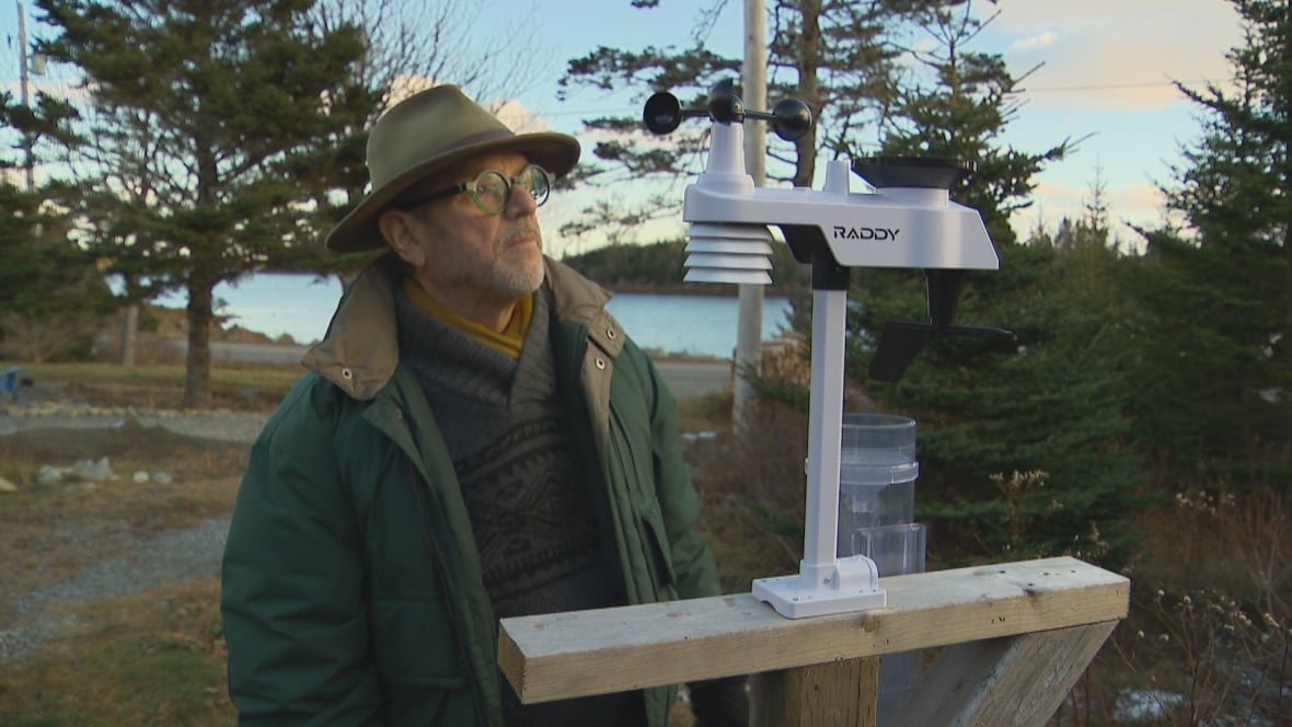 A man stands looking at an automated weather station in a wooded area with the ocean in the background.