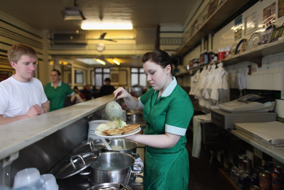 A woman in a green dress pours green parsley sauce over a plate of mashed potatoes and a minced meat pie behind the counter at a pie shop. A customer in a white tshirt waits for his plate on the other side of the counter.