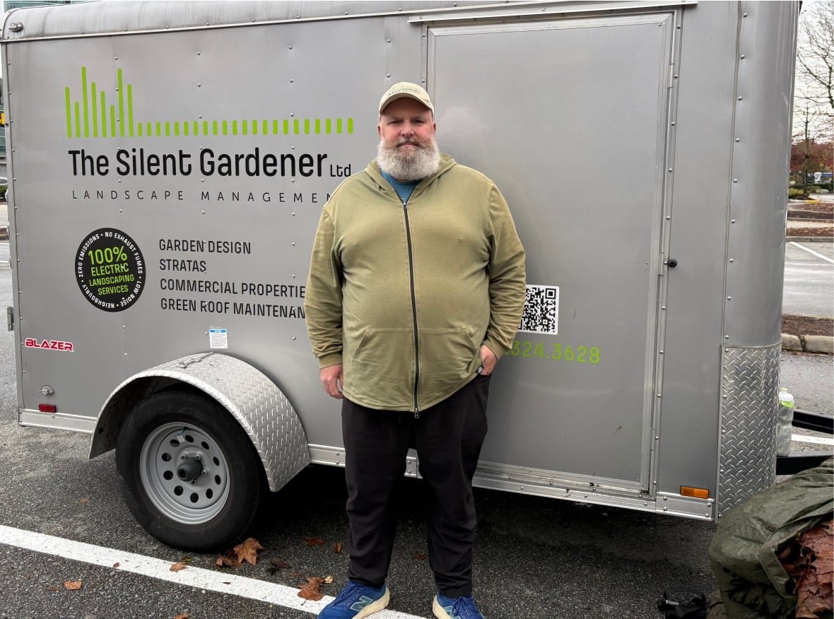 A man with a white beard wearing a white green hoodie and baseball cap stands in front of a cargo carrier that is connected to his pick-up truck.