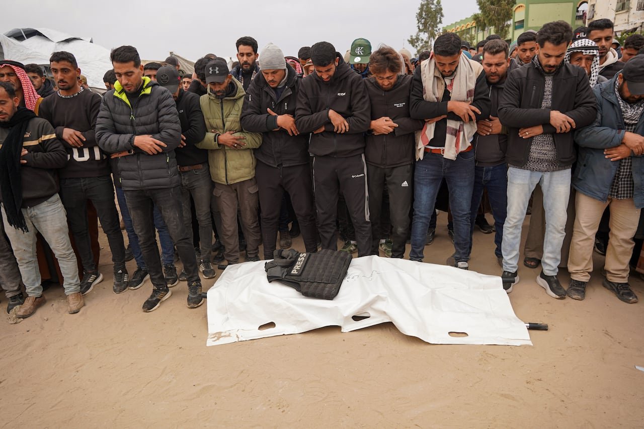 A group of individuals standing near a shrouded body with a bulletproof vest labeled 'Press' on it.