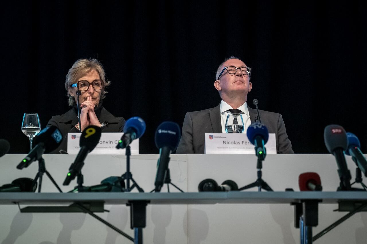 A man and a woman, both wearing glasses, sit at a long table with microphones in front. The man has tilted his head back.