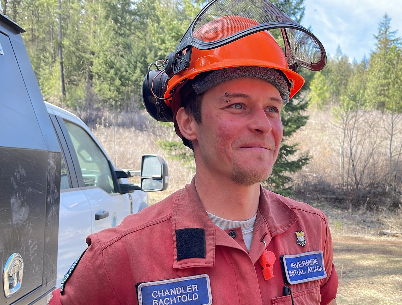 A firefighter wearing red garments and an orange helmet smiles outdoors.